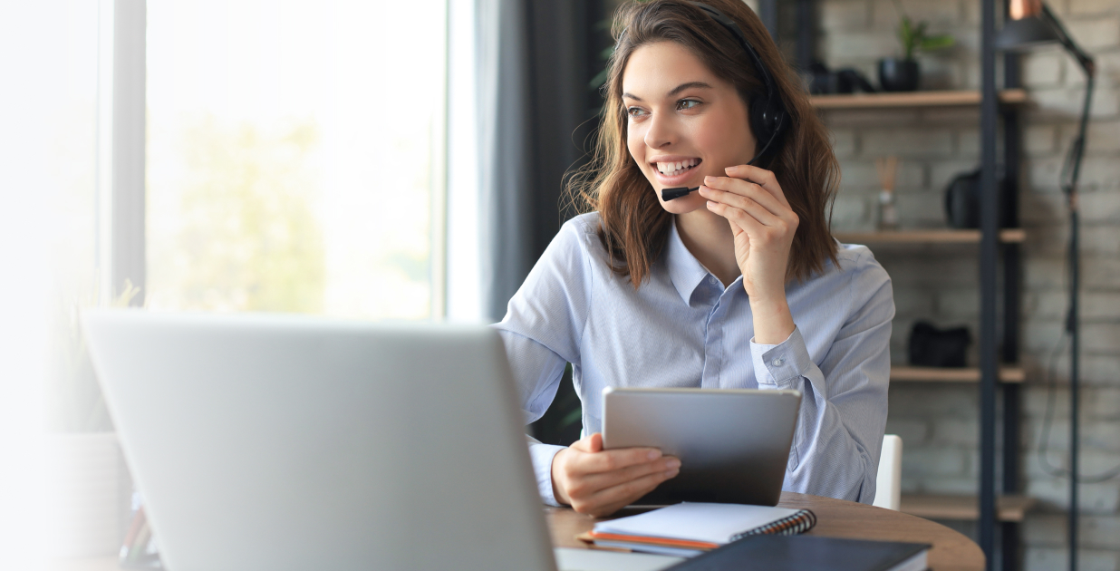 Woman smiling while using a tablet and laptop in a bright office, representing D&L Transport’s professional atmosphere for prospective freight agents